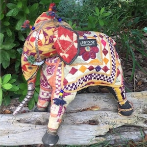 May include: A decorative elephant figurine with intricate embroidery in shades of red, yellow, orange, and purple. The elephant has a silver-colored trunk and is adorned with tassels and beads. The word "TAPOOZ" is visible on the side.