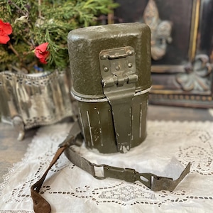 Op de afbeelding: Een olijfgroene metalen veldfles met een scharnierend deksel en een canvas riem. De veldfles heeft een rechthoekige vorm met afgeronde hoeken en een metalen sluiting. De riem is met metalen gespen aan de veldfles bevestigd. De veldfles ligt op een wit kanten kleedje.