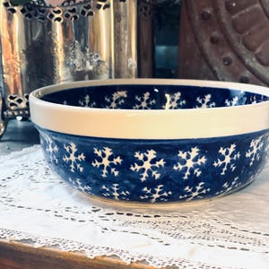 May include: A ceramic bowl with a white rim and a blue snowflake pattern. The bowl is set on a white lace doily. The bowl's interior is a deep blue, and the snowflake design is repeated around the exterior.