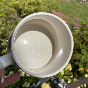 May include: A cream ceramic mug with a white handle, seen from above. The interior of the mug is smooth and reflective. The mug is set against a blurred background of green foliage and flowers.