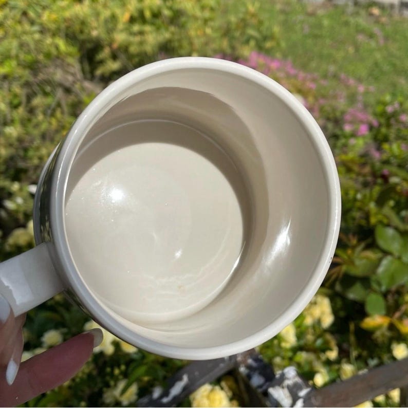 May include: A cream ceramic mug with a white handle, seen from above. The interior of the mug is smooth and reflective. The mug is set against a blurred background of green foliage and flowers.