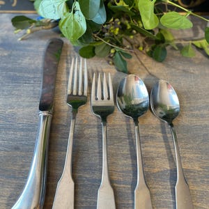 Op de afbeelding: Zilveren bestekset met een mes, twee vorken en twee lepels. Het mes heeft een gebogen handvat, terwijl de vorken en lepels eenvoudige, langwerpige handvatten hebben. Groene bladeren met bessen op de achtergrond, op een houten oppervlak.