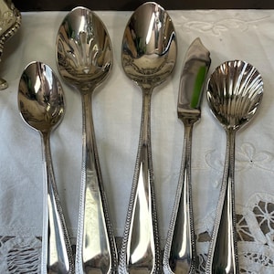 Op de afbeelding: Een verzameling van vijf zilverkleurige serveerbestekken, waaronder lepels en een botermes. De lepels hebben decoratieve, gedetailleerde kommen en handvatten. Het botermes heeft een plat, rechthoekig lemmet. De gebruiksvoorwerpen zijn gerangschikt op een wit, kantachtig oppervlak.
