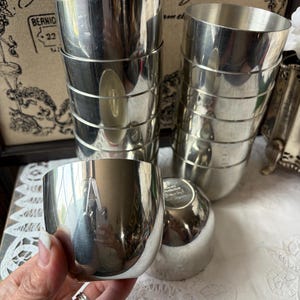 May include: A set of polished silver-toned metal cups with a smooth, reflective finish. The collection includes two tall, stacked cylindrical vessels and two shorter, rounded cups, one of which is held in the foreground to show its metallic surface.