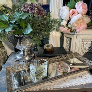 May include: A silver-toned decorative tray with ornate edges holds a rectangular perfume bottle and a collection of jewelry. The tray reflects the items and the surrounding decor, including a floral arrangement and a vintage-style table.