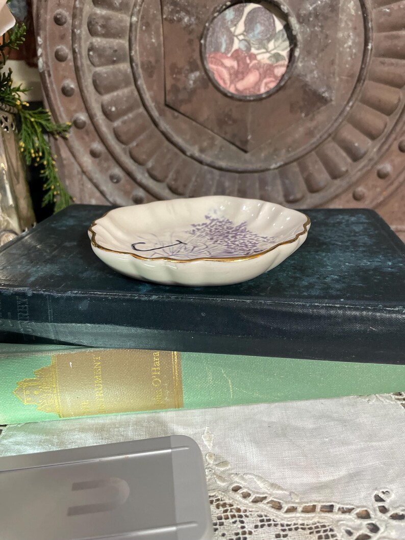 Puede incluir: Un peque&ntilde;o plato de cer&aacute;mica blanquecina con borde festoneado y borde dorado. El plato presenta un dise&ntilde;o floral morado y una l&iacute;nea negra curva. El plato est&aacute; sobre un libro azul oscuro.