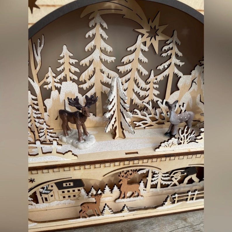 May include: A wooden Christmas scene with a winter forest design. The scene includes cut-out trees, deer, and a moose. The background features a star and a house. The overall color scheme is natural wood tones with white accents.