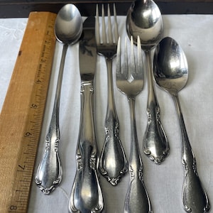 May include: A set of five silver-toned stainless steel flatware pieces including a teaspoon, a dinner knife, a dinner fork, a salad fork, and a soup spoon, all featuring ornate floral embossed handles, placed next to a wooden ruler measuring 8 inches.