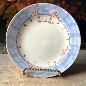 Puede incluir: Un plato de cerámica blanco con un diseño de gato pintado a mano. El gato tiene orejas y mejillas rosas, con un borde azul. El plato se exhibe en un soporte de metal dorado. Un artículo decorativo caprichoso.