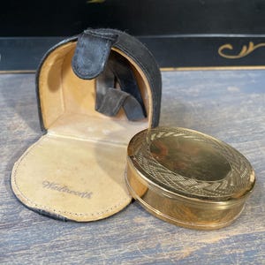 Könnte beinhalten: Eine offene schwarze Ledertasche mit einem goldfarbenen ovalen Kompakt daneben. Das Kompakt hat ein eingraviertes Design auf dem Deckel. Die Tasche ist mit beigem Stoff gefüttert, und das Wort "Wadsworth" ist auf den Stoff gedruckt.