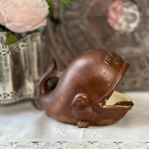 以下が含まれることがあります： 茶色のセラミック製のクジラの置物。口が開いており、クリーム色の内側が見える。クジラは笑顔で、白いレースのトリムが付いた表面に置かれている。「FOR A OF」の文字が上部に書かれている。