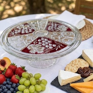 Pode incluir: Um suporte para bolos em vidro transparente com um topo circular que apresenta painéis alternados de vidro prensado transparente e vidro vermelho profundo. As secções vermelhas são decoradas com cachos de uvas e folhas gravadas.