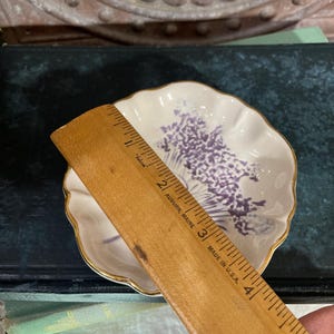 Puede incluir: Un peque&ntilde;o plato blanco con borde festoneado, borde dorado y dise&ntilde;o floral morado. El plato se mide con una regla de madera. La regla est&aacute; marcada en cent&iacute;metros.