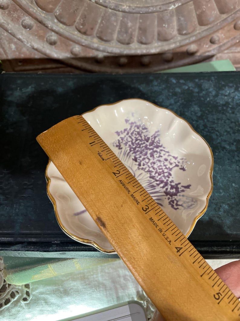 Puede incluir: Un peque&ntilde;o plato blanco con borde festoneado, borde dorado y dise&ntilde;o floral morado. El plato se mide con una regla de madera. La regla est&aacute; marcada en cent&iacute;metros.