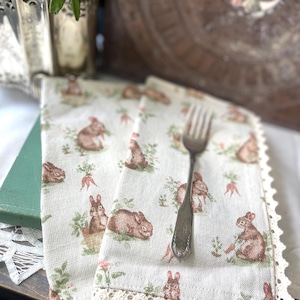 Può includere: Due tovaglioli di lino bianco con un motivo ripetuto di conigli marroni, carote e fogliame verde. I tovaglioli hanno un bordo in pizzo color crema e una forchetta d'argento è appoggiata sopra. I tovaglioli sono su un tavolo con un libro verde e una composizione floreale.