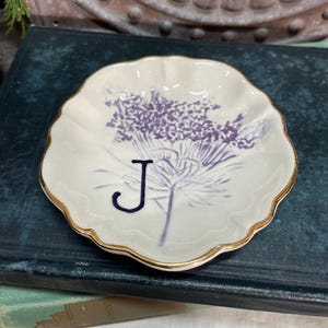 Puede incluir: Un peque&ntilde;o plato de cer&aacute;mica blanco con borde festoneado y ribete dorado. El plato presenta un dise&ntilde;o floral morado y la letra "J" azul marino. Este plato decorativo es ideal para guardar objetos peque&ntilde;os.