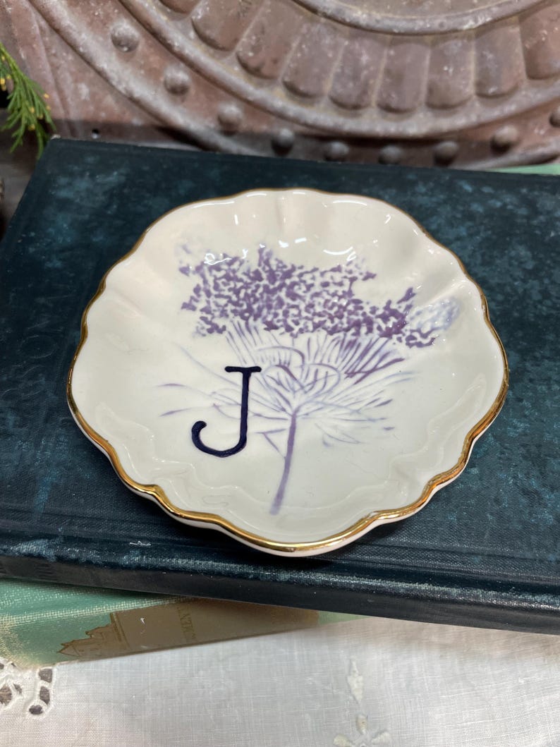 Puede incluir: Un peque&ntilde;o plato de cer&aacute;mica blanco con borde festoneado y ribete dorado. El plato presenta un dise&ntilde;o floral morado y la letra "J" azul marino. Este plato decorativo es ideal para guardar objetos peque&ntilde;os.