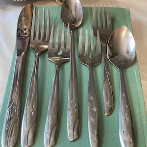 Puede incluir: Un conjunto de cubiertos antiguos, que incluye tenedores, cucharas y un cuchillo, dispuestos sobre una superficie verde claro. La cubertería presenta un diseño de estrella en los mangos. El cuchillo está a la izquierda.