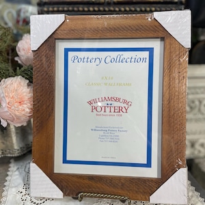 May include: A wooden 8x10 inch classic wall frame from the Pottery Collection. The frame has a brown, textured finish and is wrapped in protective plastic. The frame includes the text "Williamsburg Pottery" and "Best buys since 1938".