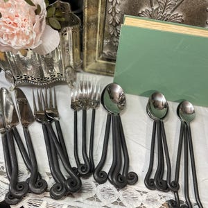 May include: A collection of silverware, including knives, forks, and spoons, with dark metal handles featuring a decorative scroll design. The silverware is arranged on a white lace tablecloth, suggesting a vintage or rustic aesthetic.