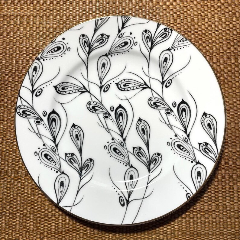 May include: White ceramic plate with a black and white floral design. The plate features a gold rim and a repeating pattern of stylized leaves and teardrop shapes. The design is intricate and detailed.