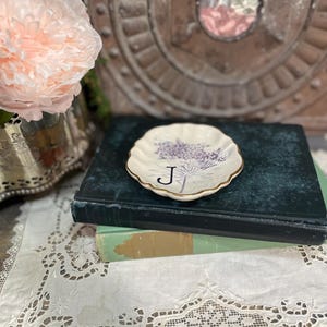 Puede incluir: Un plato peque&ntilde;o, blanco, con borde festoneado y borde dorado, con un dise&ntilde;o de ramita de lavanda y la letra "J" en negro. El plato descansa sobre una pila de libros, con un fondo floral vintage.