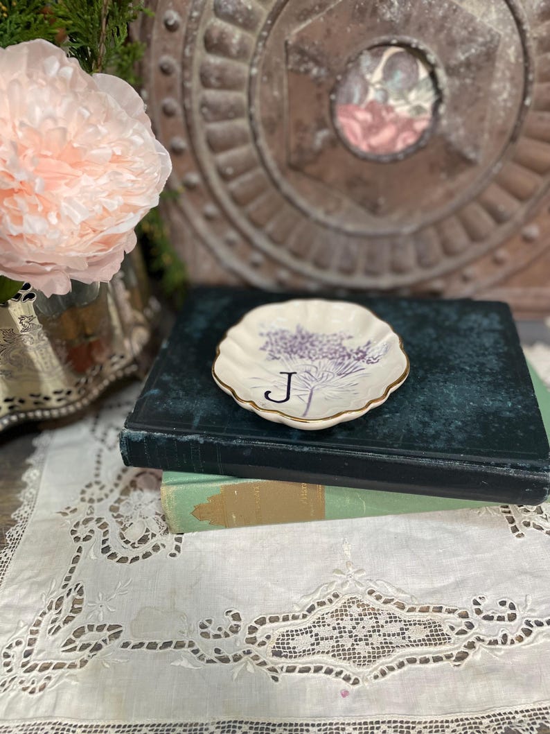 Puede incluir: Un plato peque&ntilde;o, blanco, con borde festoneado y borde dorado, con un dise&ntilde;o de ramita de lavanda y la letra "J" en negro. El plato descansa sobre una pila de libros, con un fondo floral vintage.