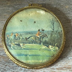 May include: A round, antique compact with a hinged lid and a gold-toned metal frame. The interior features a painted scene of a fox hunt with riders on horseback, hounds, and a green landscape.