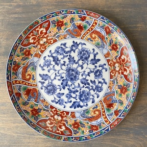 Puede incluir: Un plato decorativo de cerámica con un diseño floral. El plato presenta un centro blanco con flores azules, rodeado por un borde de patrones rojos, amarillos y azules. El plato es redondo y tiene un acabado brillante.
