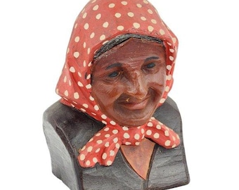 May include: A carved wooden bust of an elderly woman. The woman has dark skin, a smiling expression, and is wearing a red headscarf with white polka dots. Her clothing is dark, and the bust is set against a white background.