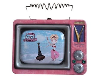 Puede incluir: Una lata de colección de metal rosa con forma de televisor antiguo. La pantalla presenta un gráfico de I Dream of Jeannie con un genio y una botella. La lata incluye una antena de metal, un logotipo de Vandor, dos perillas de control y dos botones verdes.