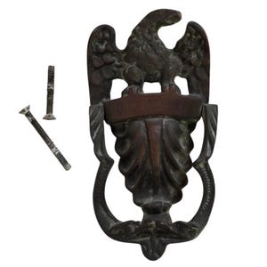 May include: A dark bronze-toned metal door knocker featuring an eagle at the top and a central shield-like element with a horizontal banner inscribed with the text O CHIAVETTA. Two metal mounting screws are positioned to the left of the knocker.