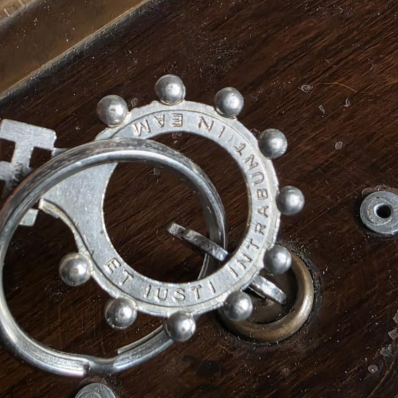 Pode incluir: Close-up de um artefato religioso de metal prateado com a inscri&ccedil;&atilde;o "ET IUSTI IN TRAMEAM". O artefato tem um design circular com pequenas decora&ccedil;&otilde;es redondas e est&aacute; sobre uma superf&iacute;cie de madeira escura.