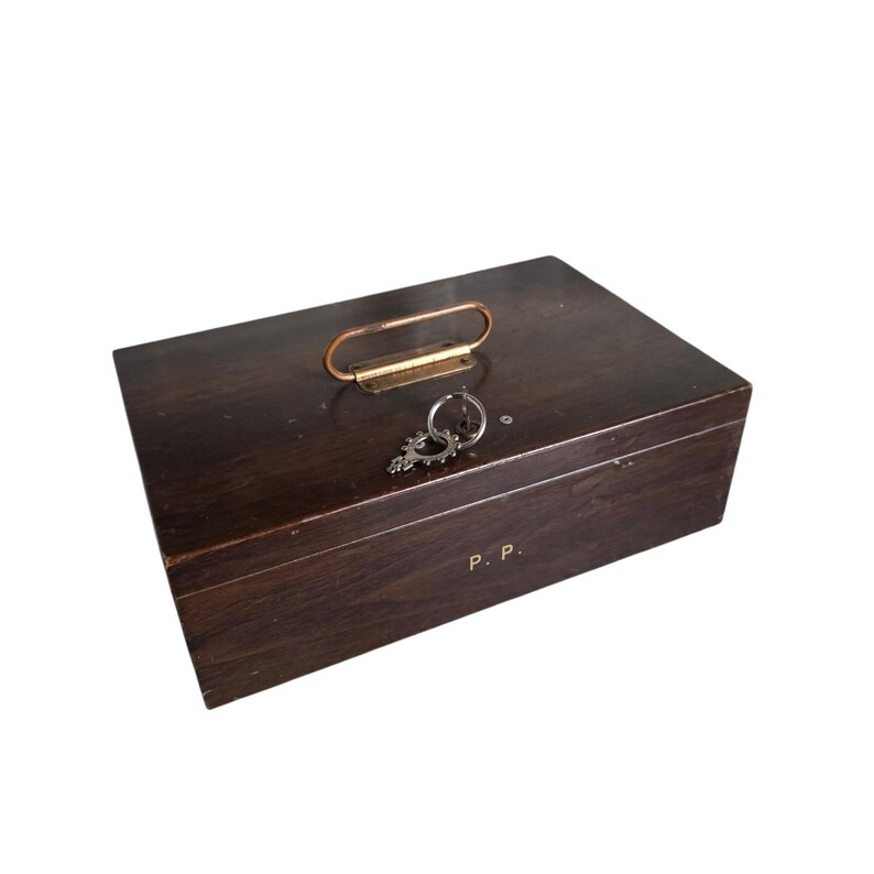 Pode incluir: Uma caixa de madeira castanha escura com uma pega de metal e uma chave. A caixa tem as iniciais "P. P." na frente. A pega &eacute; de cor cobre. A chave est&aacute; presa a um anel.