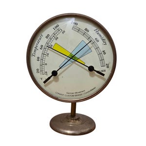 Op de afbeelding: Een ronde Conant Custom Brass ComfortMeter van messing met een witte wijzerplaat, twee zwarte wijzers, een temperatuurschaal in Fahrenheit, een vochtigheidsschaal in procenten en een centrale gele en blauwe comfortzone op een ronde voet.