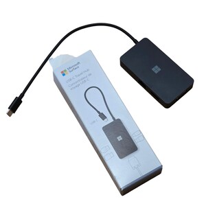 May include: A Microsoft Surface USB-C Travel Hub with a black cable and a black rectangular hub. The box is white with the Microsoft logo and product information. The text "USB-C Travel Hub" is visible on the box.