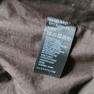 May include: A black tag from AllSaints Spitalfields with text detailing the outer and inner materials of a Vancouver Jacket. The tag includes the RN number, fabric composition, and manufacturing location.