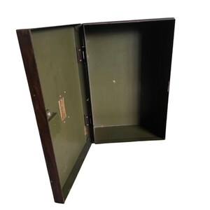 Pode incluir: Uma caixa de metal aberta, verde-oliva, com uma moldura castanha escura. A caixa tem formato retangular com uma tampa articulada. Uma pequena placa de metal retangular com texto &eacute; vis&iacute;vel na frente. O interior &eacute; verde-oliva.