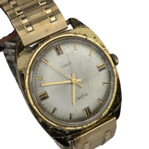 Könnte beinhalten: Vintage goldfarbene Timex Electric Uhr mit silbernem Zifferblatt. Die Uhr hat goldene Zeiger und Stundenmarkierungen sowie ein goldfarbenes Metallarmband. Das Wort "TIME" steht über der Mitte und "Electric" darunter.