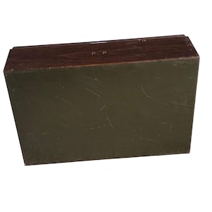 Pode incluir: Uma caixa retangular de madeira com uma parte superior castanha escura e laterais verde-oliva. A parte superior da caixa tem as iniciais "P. P." numa cor mais clara. A caixa parece ser vintage ou antiga.