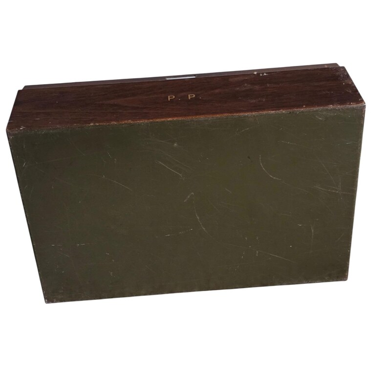 Pode incluir: Uma caixa retangular de madeira com uma parte superior castanha escura e laterais verde-oliva. A parte superior da caixa tem as iniciais "P. P." numa cor mais clara. A caixa parece ser vintage ou antiga.