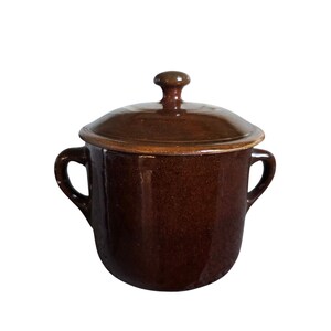 May include: A dark brown glazed ceramic pot with two side handles and a matching lid featuring a central knob. The vessel has a cylindrical body with a smooth, reflective finish and a slightly flared rim, presenting a uniform deep brown color throughout.