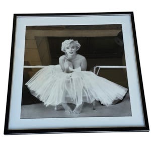 Może przedstawiać: Czarno-biała oprawiona fotografia Marilyn Monroe w białej tiulowej sukni. Zdjęcie przedstawia Monroe siedzącą, ze splecionymi dłońmi blisko piersi. Fotografia jest w czarnej ramie z białym passe-partout.