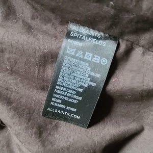 May include: A black label with white text from AllSaints Spitalfields. The label includes the RN number, fabric composition, and the text "Vancouver Jacket". The label is on a brown fabric background.