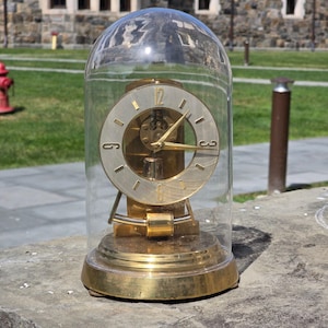 May include: A gold-toned anniversary clock with a circular brushed metal face, black numerals, and gold hands, enclosed within a clear glass dome. The clock features a brass base and a visible internal pendulum mechanism behind the clock face.