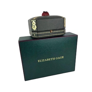 May include: A dark green jewelry box with gold trim and a small, matching top box. The top box has a red tassel and gold detailing. The larger box has the name "ELIZABETH GAGE" printed in gold.