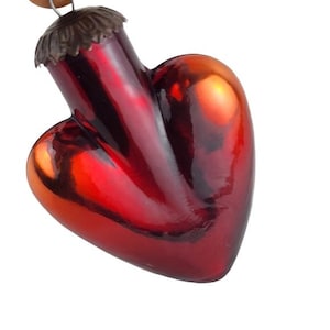 Puede incluir: Un adorno navideño en forma de corazón con un acabado rojo vibrante. El adorno tiene una superficie brillante y reflectante y una tapa de metal marrón. El diseño de corazón es una decoración navideña clásica.