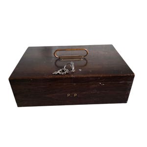 Pode incluir: Uma caixa de madeira castanha escura com uma pega de metal e uma chave. A caixa tem formato retangular e um aspeto ligeiramente desgastado. A chave &eacute; prateada e ornamentada. As iniciais "P. P." s&atilde;o vis&iacute;veis na frente da caixa.