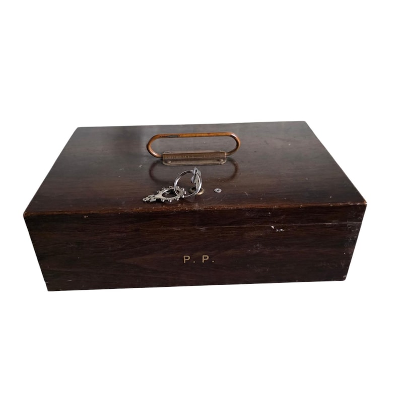 Pode incluir: Uma caixa de madeira castanha escura com uma pega de metal e uma chave. A caixa tem formato retangular e um aspeto ligeiramente desgastado. A chave &eacute; prateada e ornamentada. As iniciais "P. P." s&atilde;o vis&iacute;veis na frente da caixa.