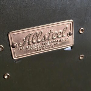 Pode incluir: Close-up de uma placa de metal bronze com a palavra "Allsteel" em uma fonte elegante. A placa diz "THE GENERAL FIREPROOFING CO. YOUNGSTOWN, OHIO. MADE IN U.S.A." e &eacute; fixada a uma superf&iacute;cie verde escura com rebites.